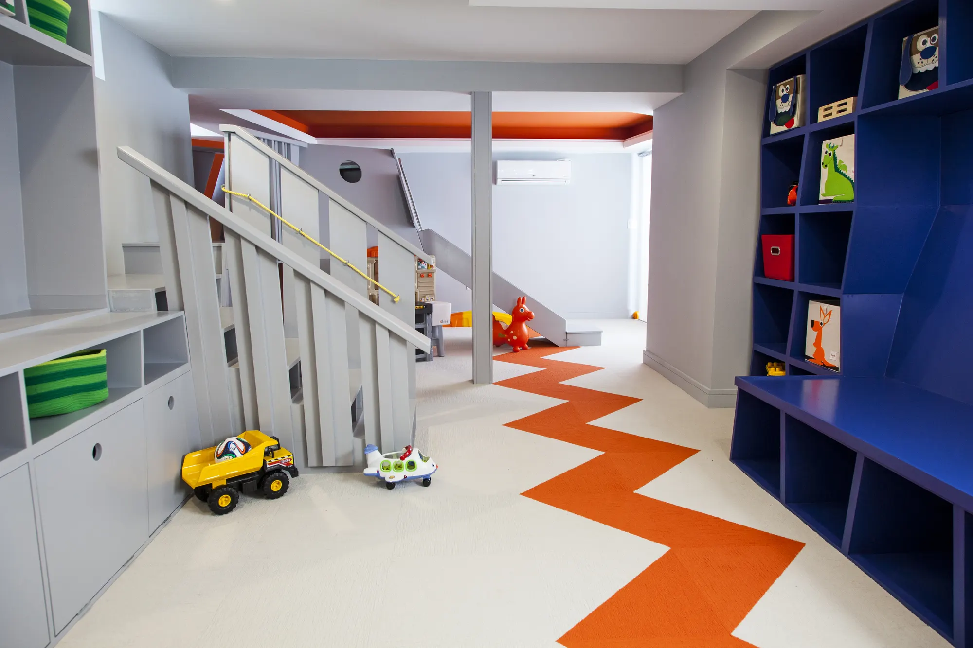 The grey stair, blue cubbies and orange zigzag floor, with toy trucks in front.