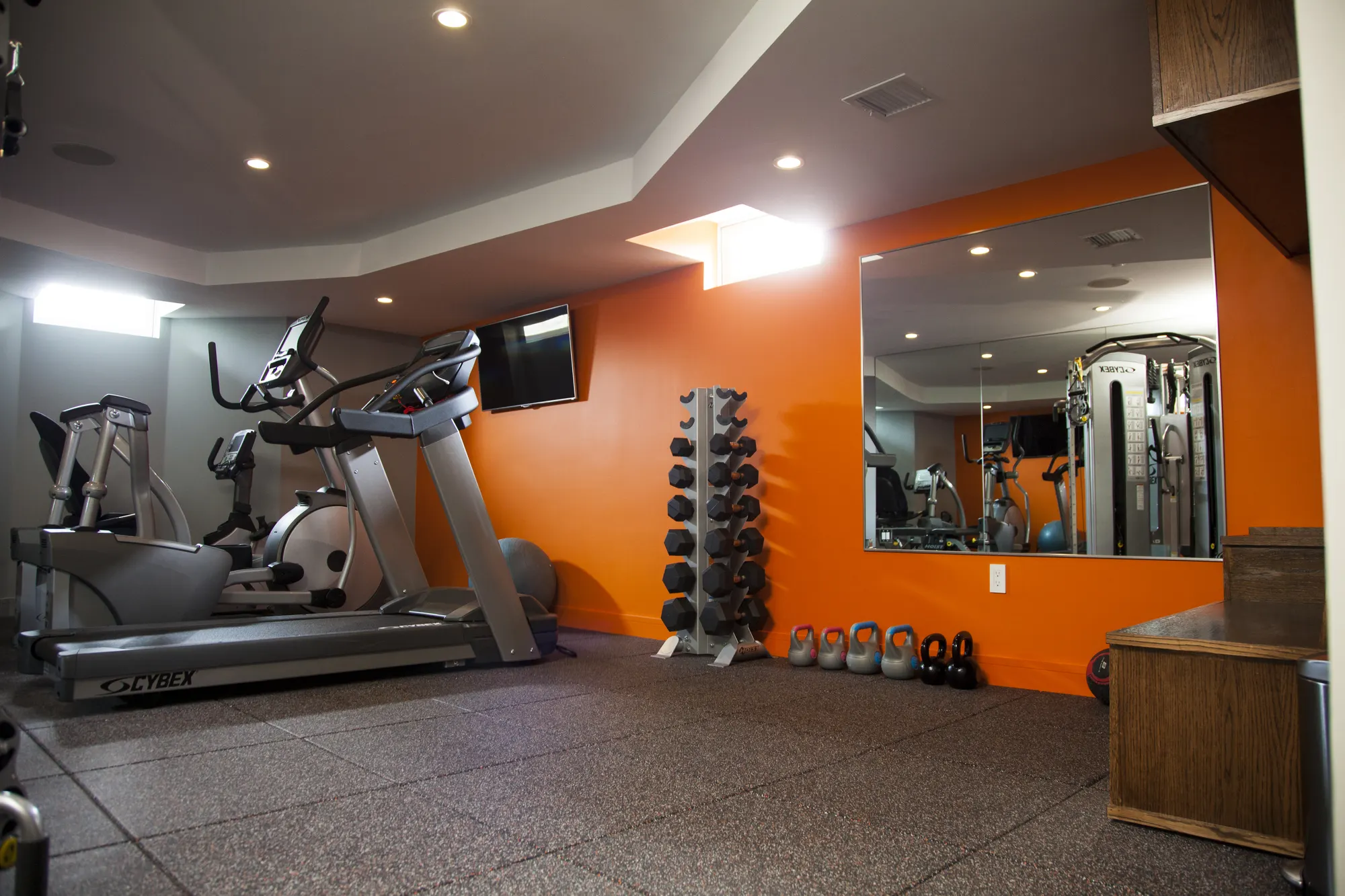 A treadmill, an exercise bike and a rack of dumbbells against an orange wall with a mirror.