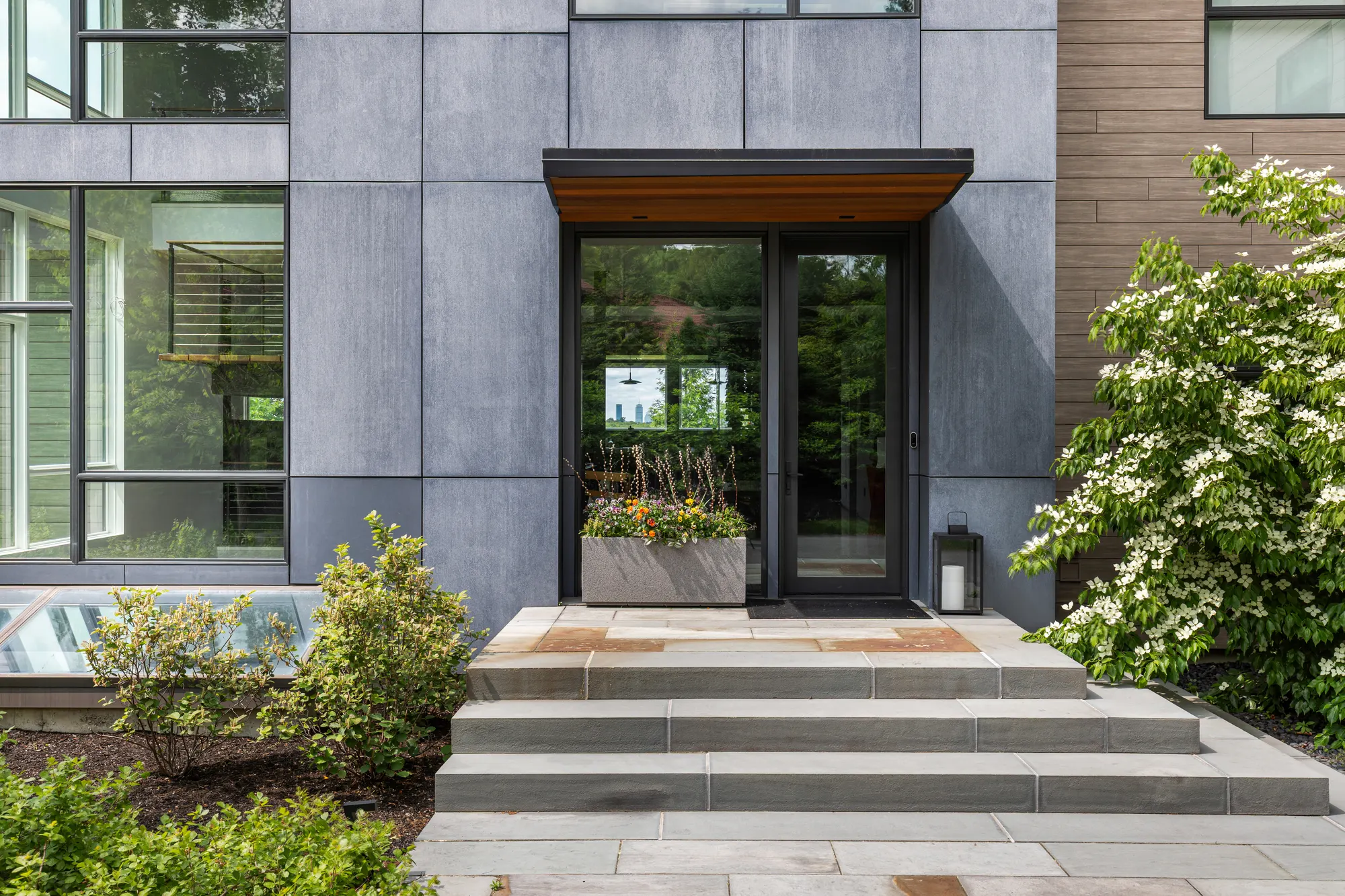 Broad stone steps up to a glazed front door under a timber-lined canopy, set in grey panelled cladding.