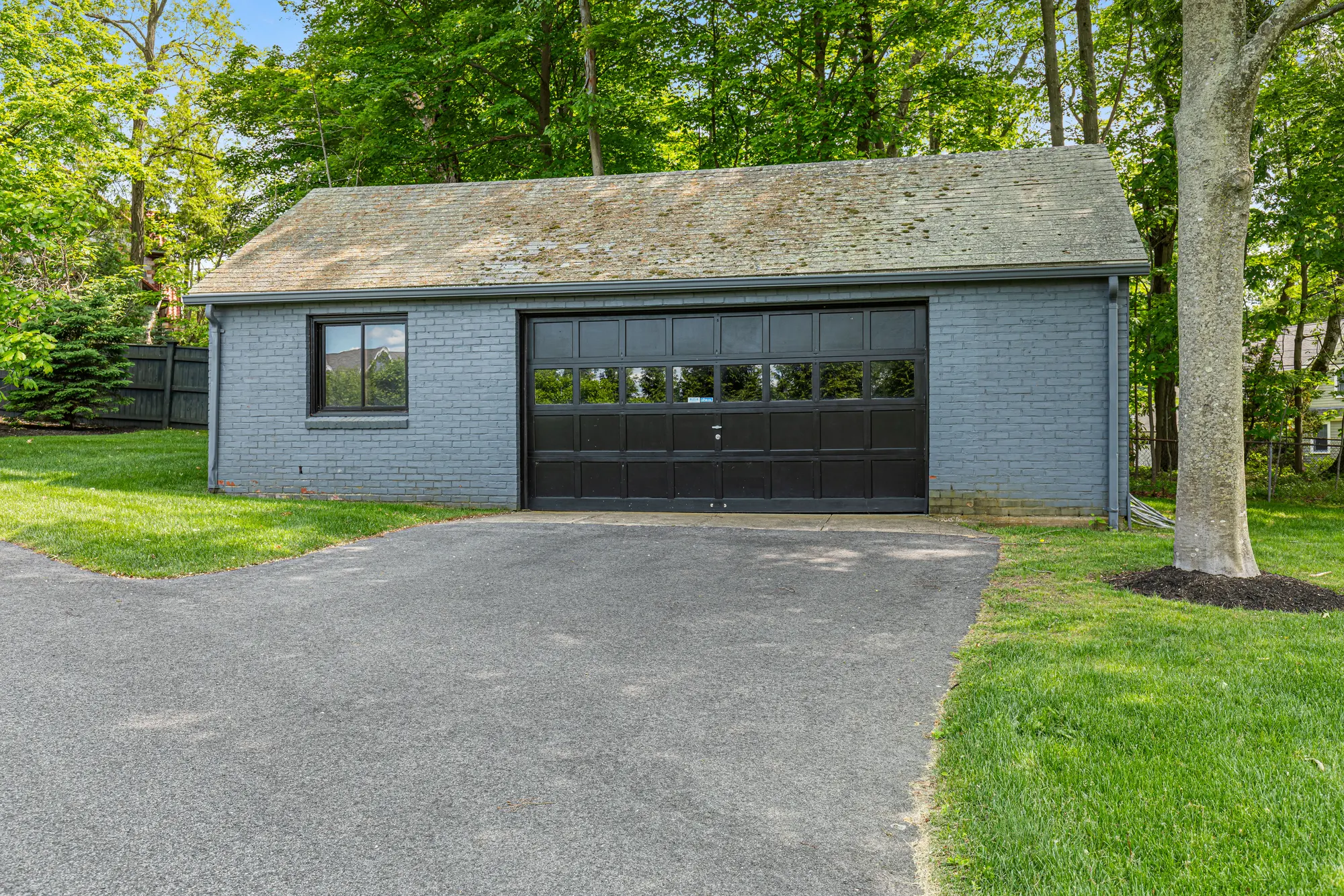 A grey brick garage with a dark door at the end of the drive, among trees.