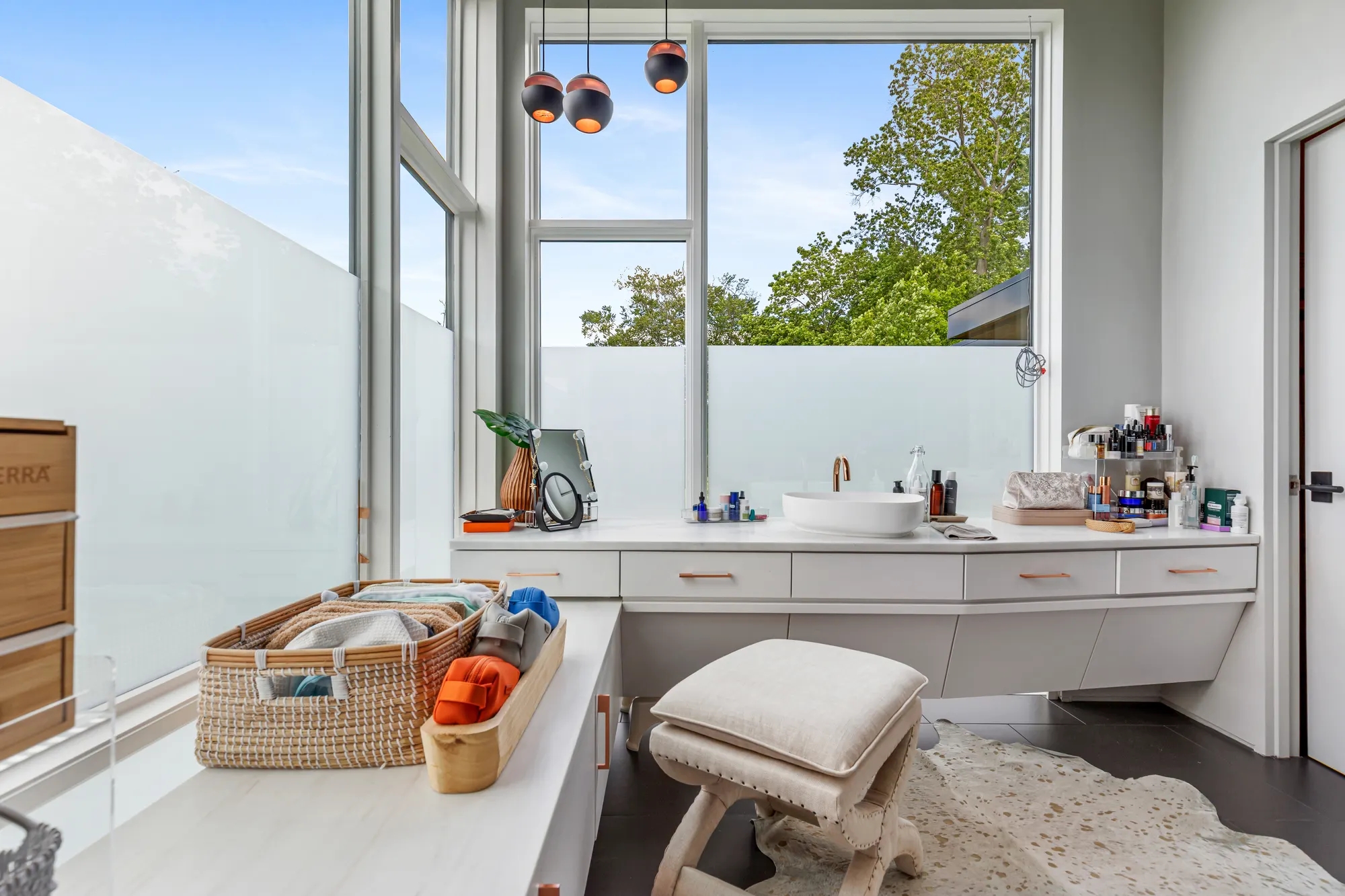 The vanity and a stool under a tall corner window, a basket of towels on the counter.