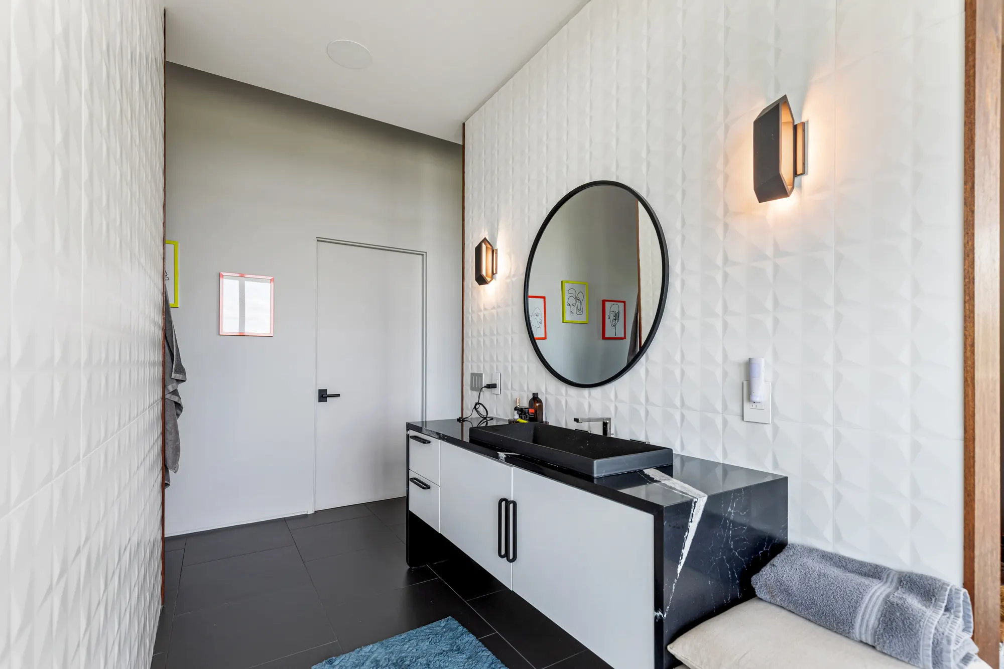 A black vanity with a round mirror and wall light, against white faceted tile.