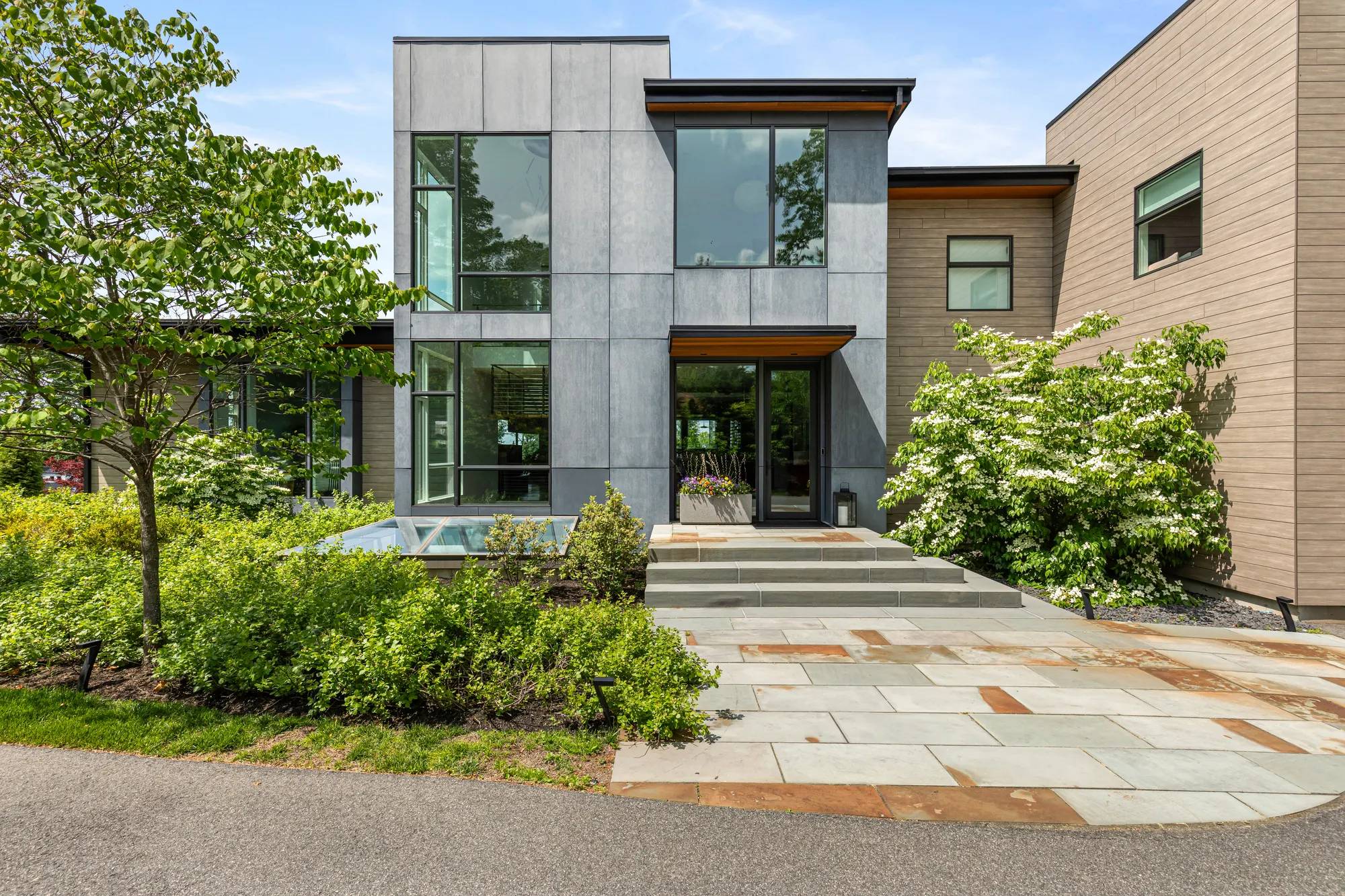 The grey panelled entry block and its front door, reached by stone paving and steps through planting.