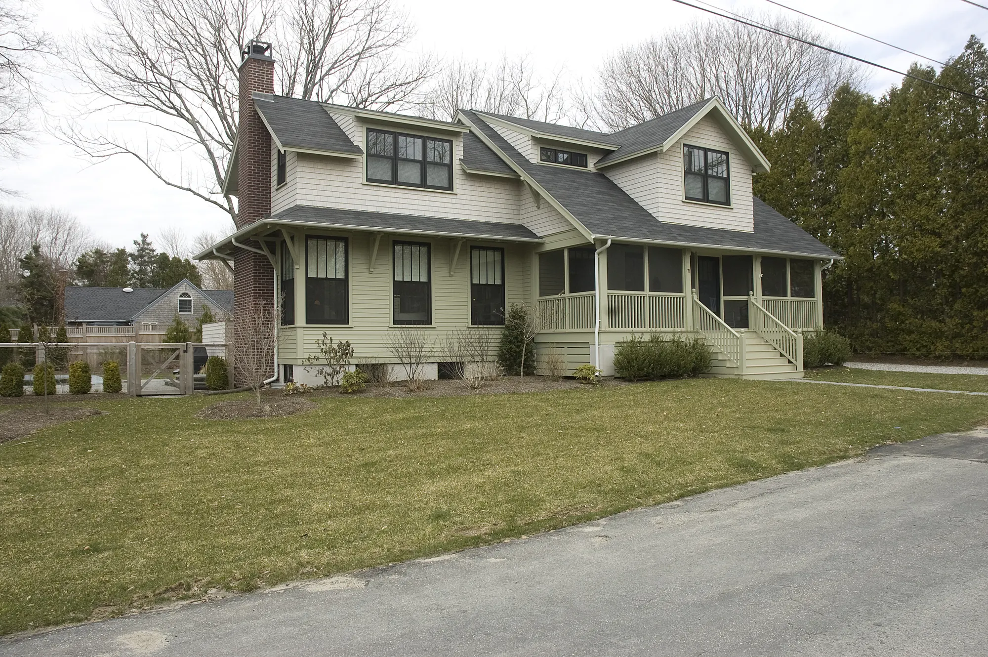 A shingled house with a deep front porch and dormers, set back from the road behind a lawn.