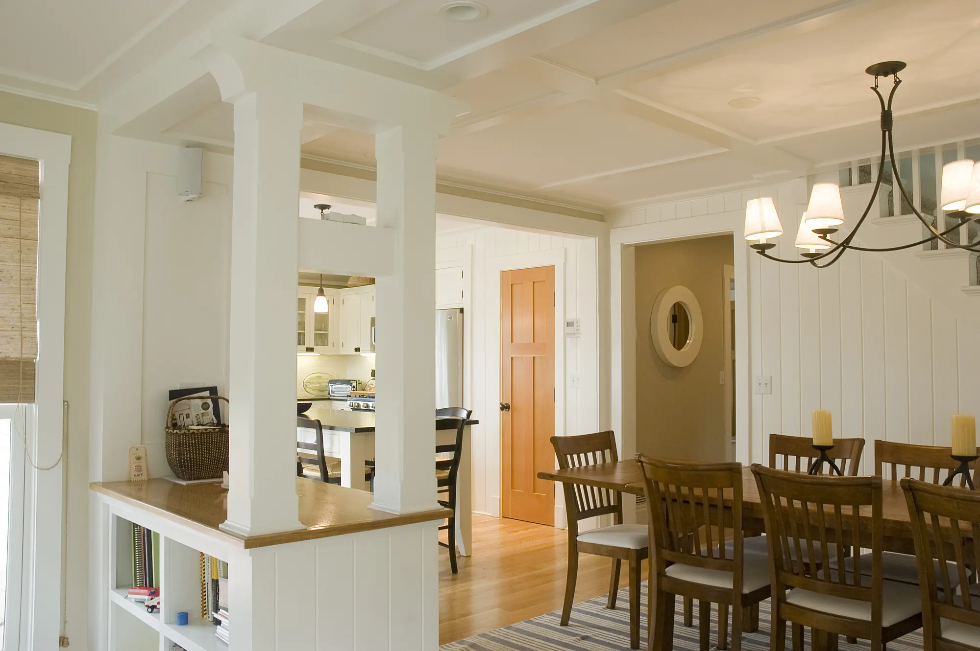 A timber dining table under a chandelier between white columns, with the kitchen beyond.