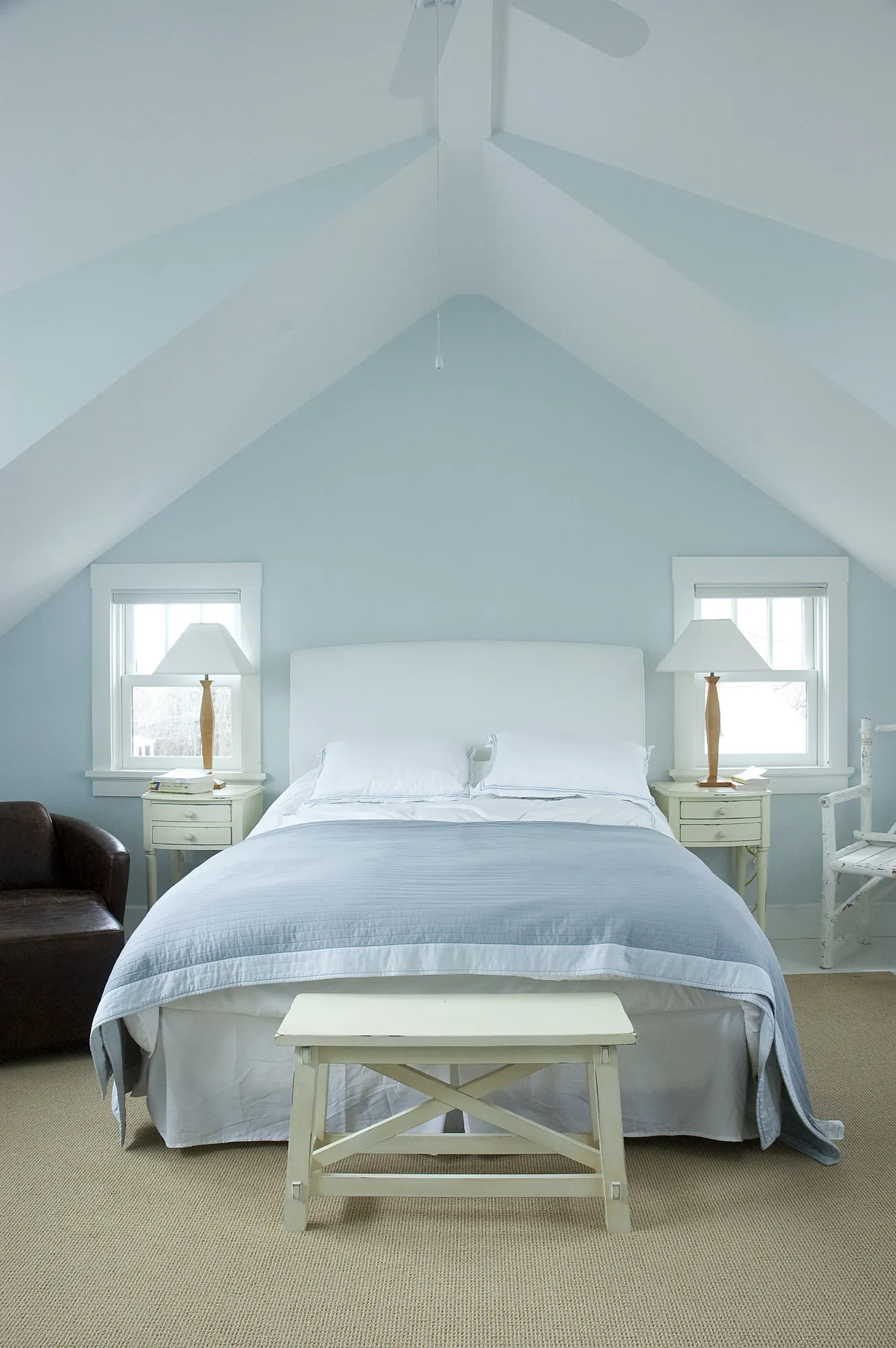 A white bed under a pitched ceiling painted pale blue, between two small windows.