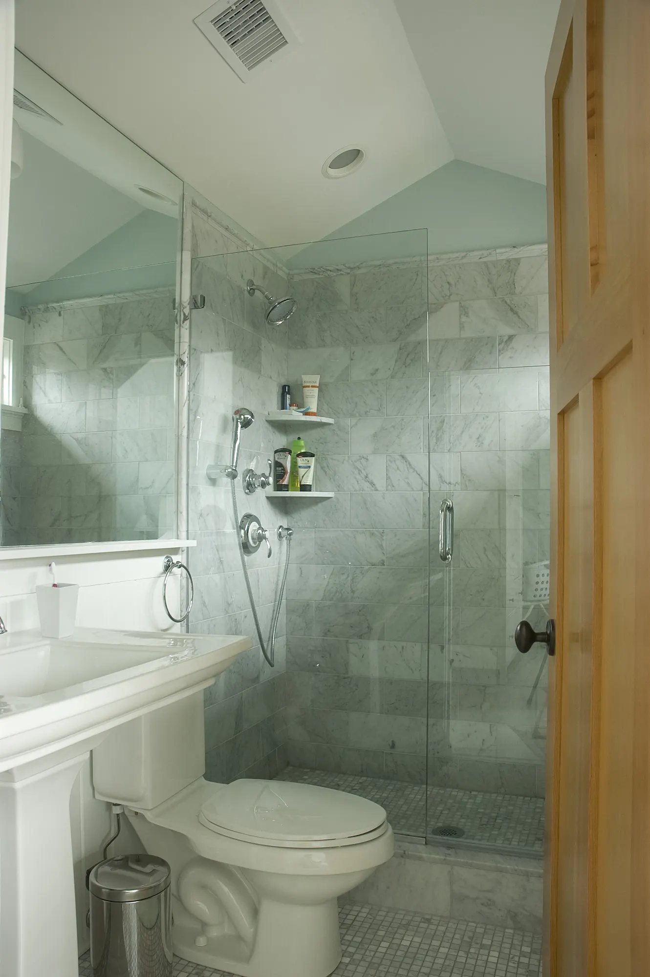A glass shower in grey marble beside a pedestal basin, under a sloped ceiling.