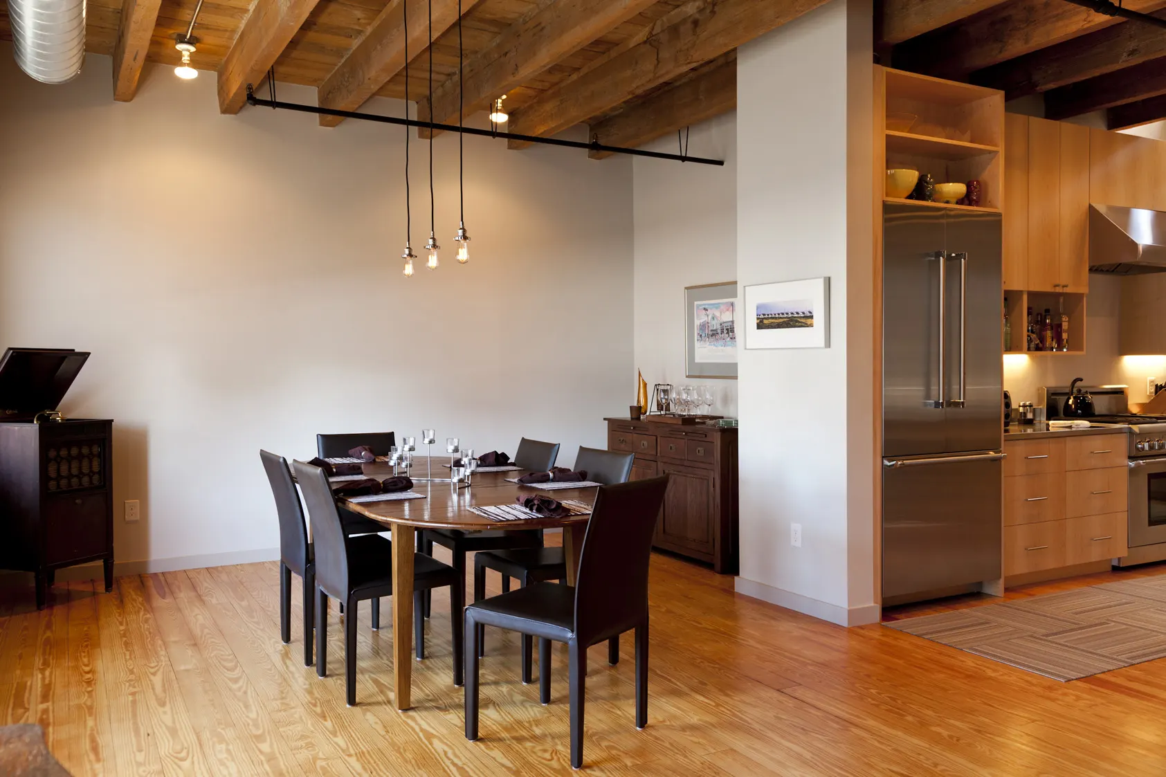 A dining table under timber beams and pendant lights, with the maple kitchen and a steel fridge beyond.