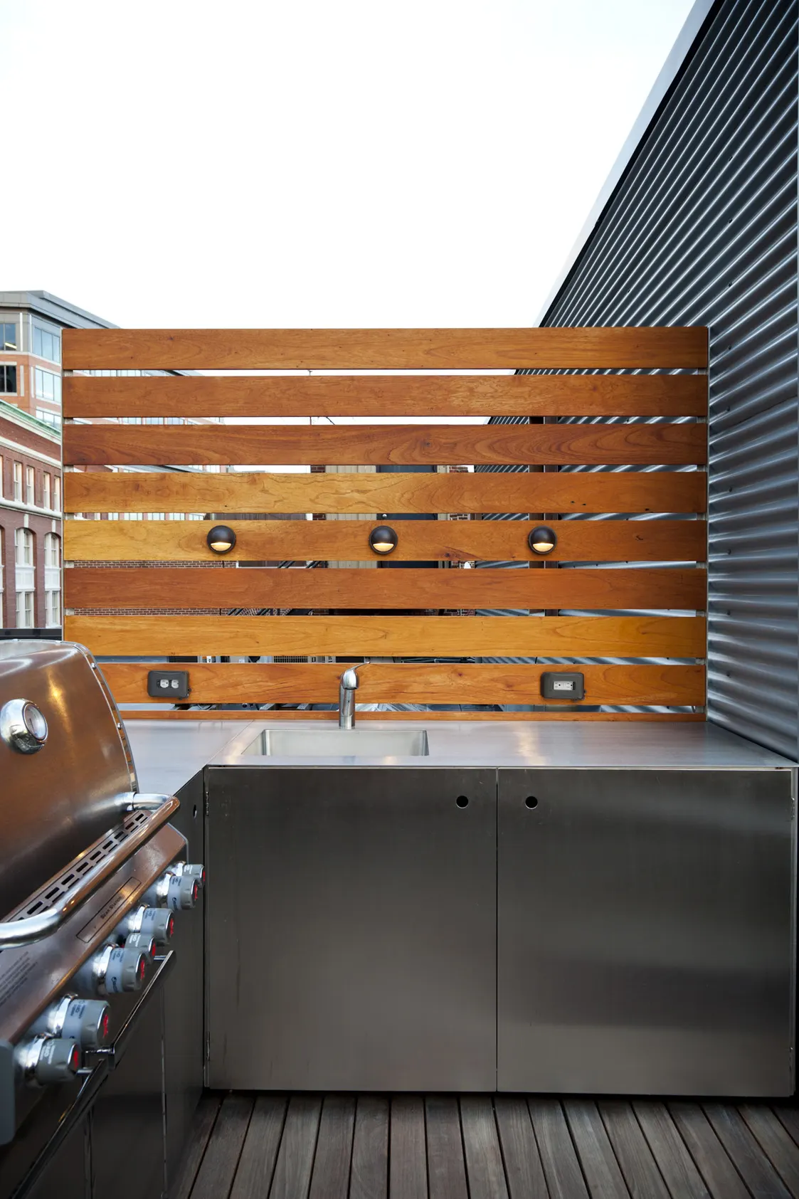 A grill and a stainless steel counter with a sink, against a slatted timber screen.