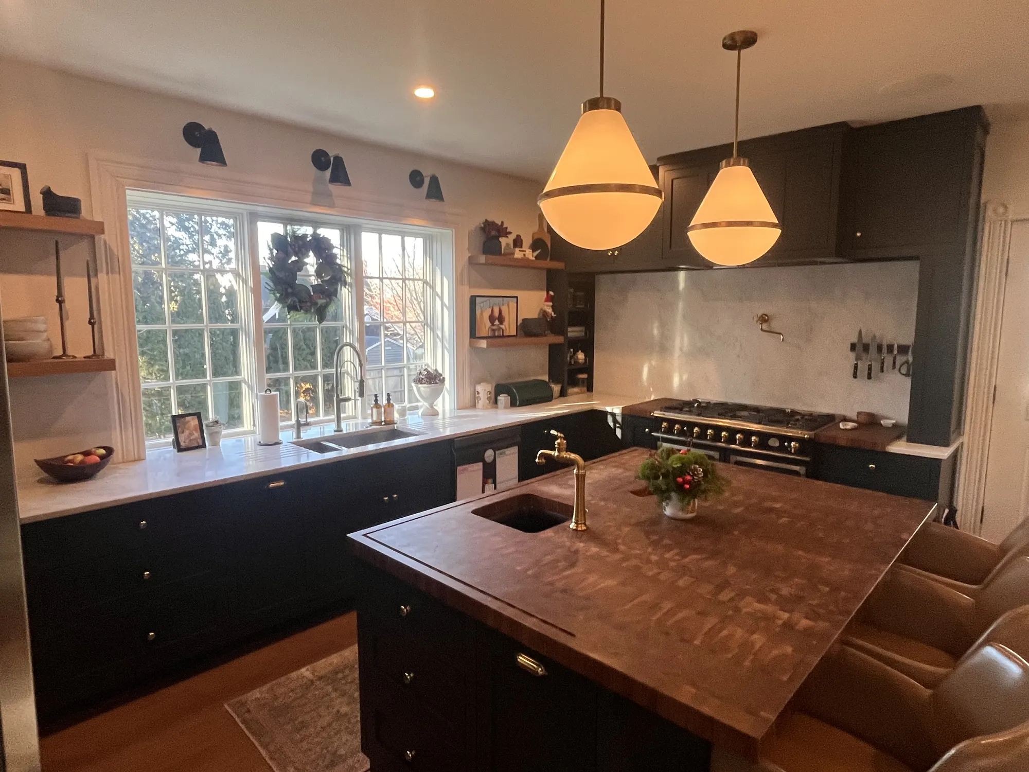 The sink under a wide window hung with a wreath, open shelves either side, and the walnut island in front.