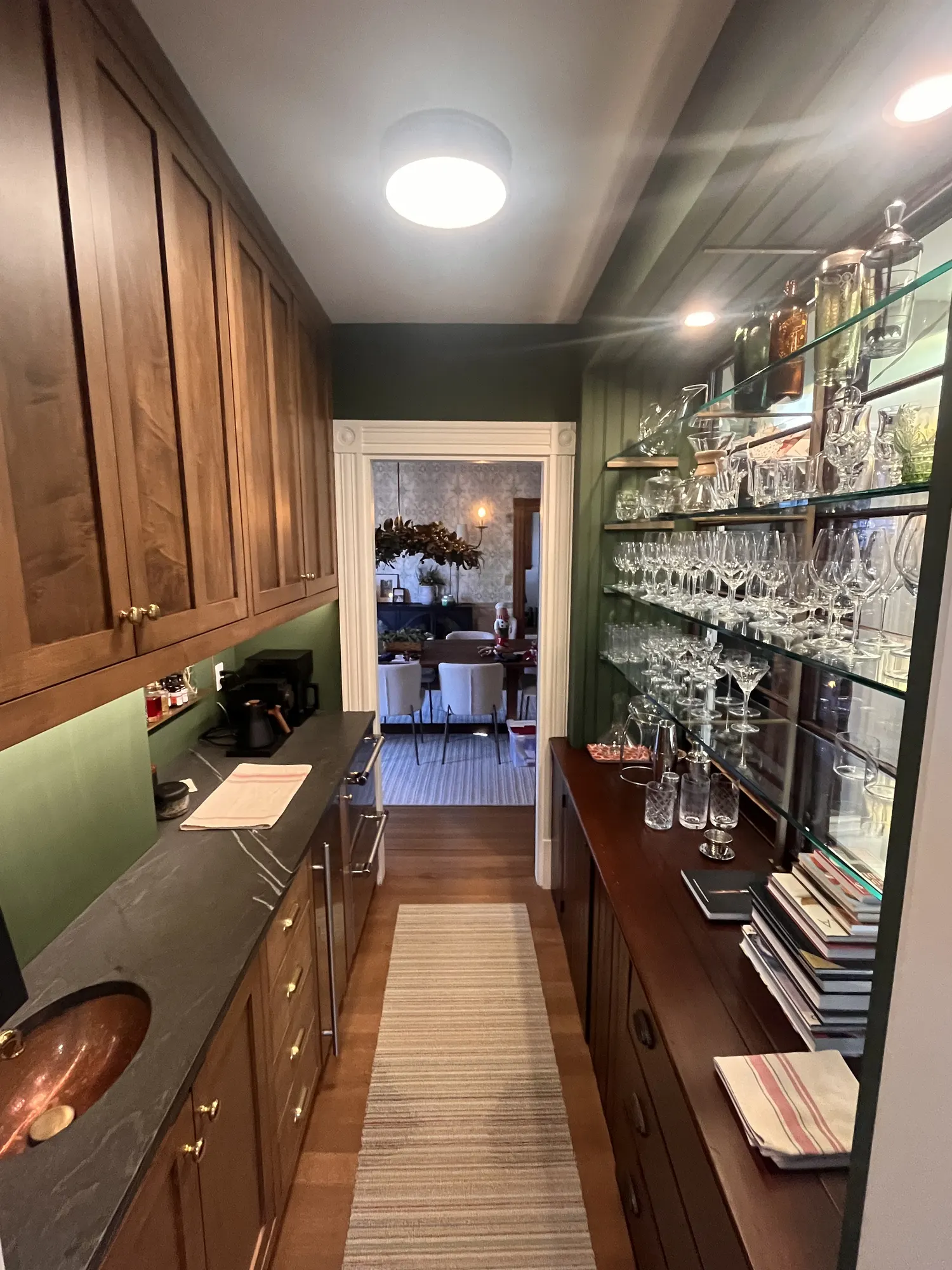 A narrow pantry with green walls: walnut cabinets and a copper sink on one side, glass shelves of glassware on the other.