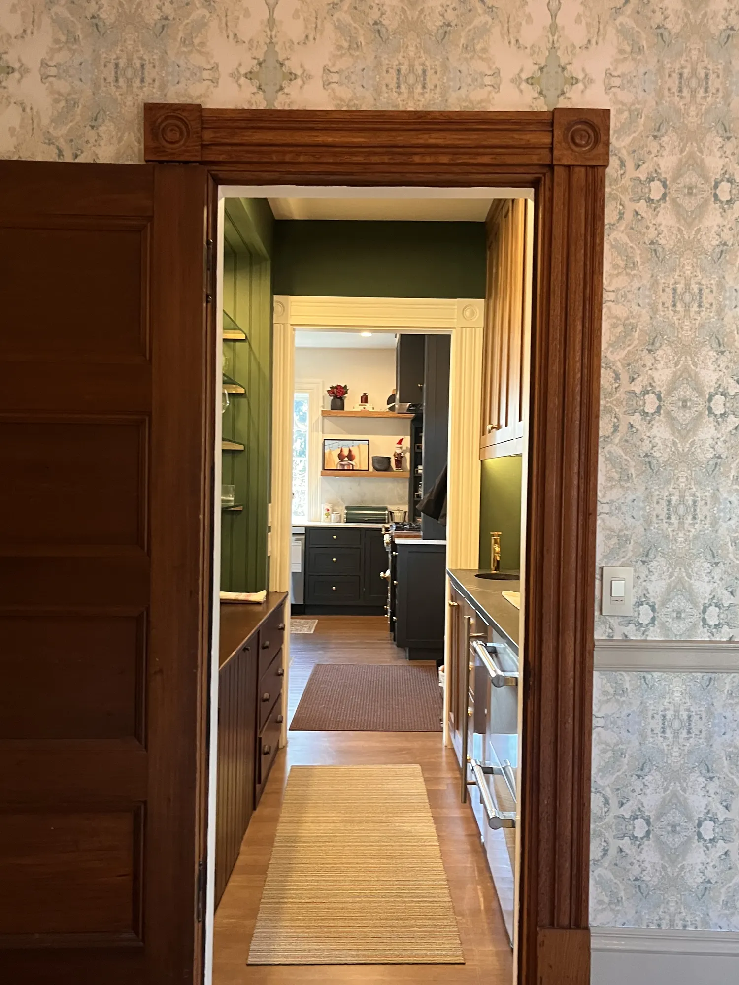 A dark timber doorway in a patterned wallpaper wall, looking through the pantry to the kitchen.
