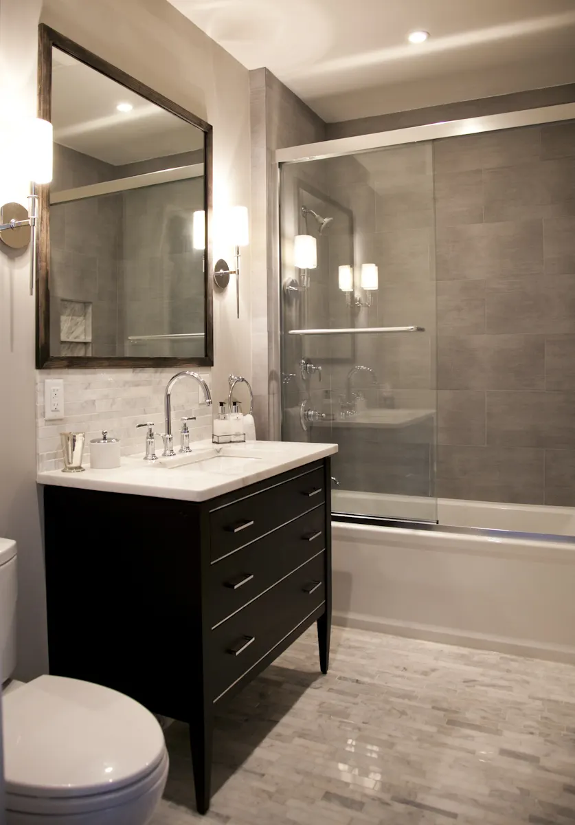 A black vanity with a white top under a large mirror, beside a glass-screened bath in grey tile.