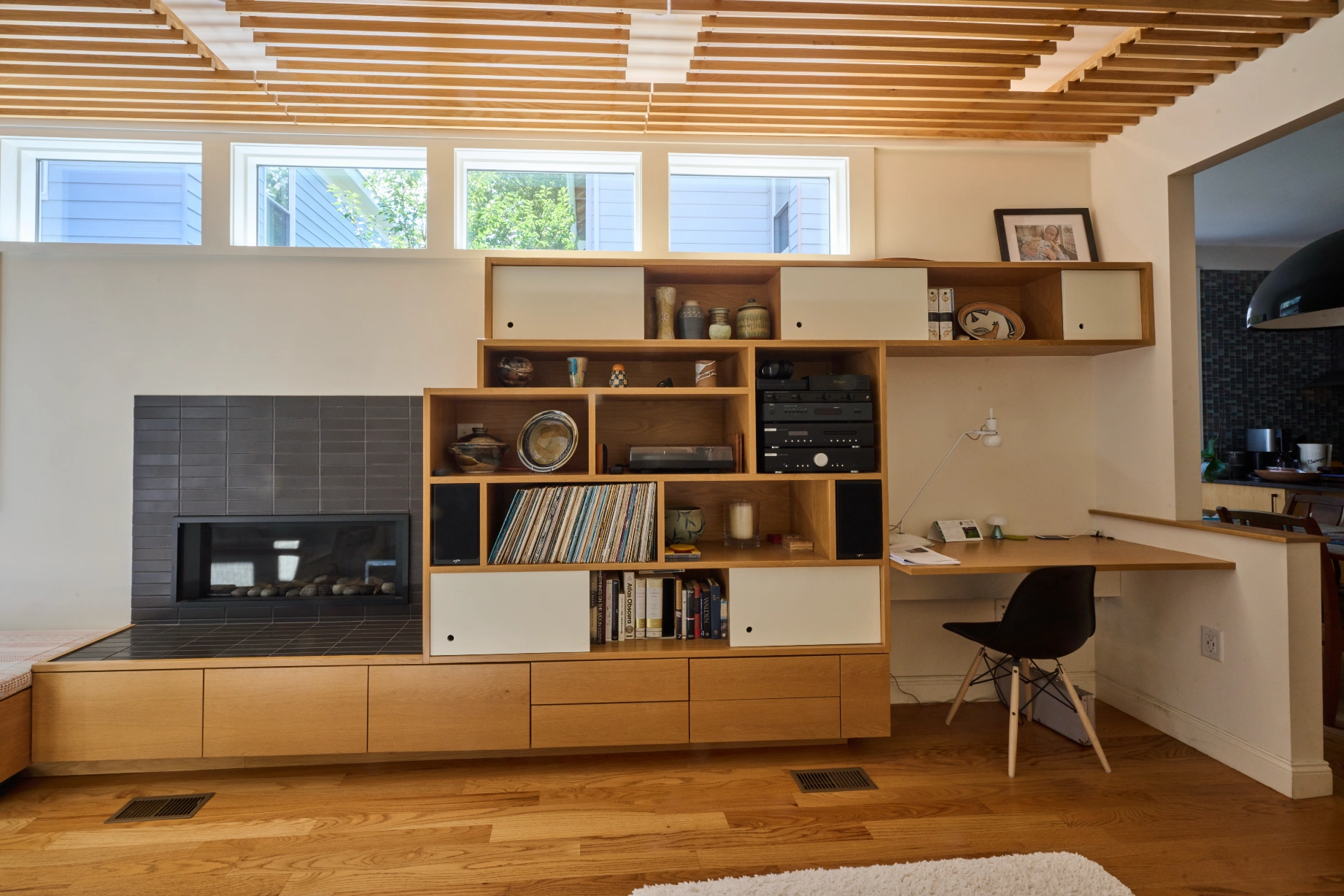 The maple built-in wall with the fireplace, shelving and a desk and chair, under the slatted ceiling.