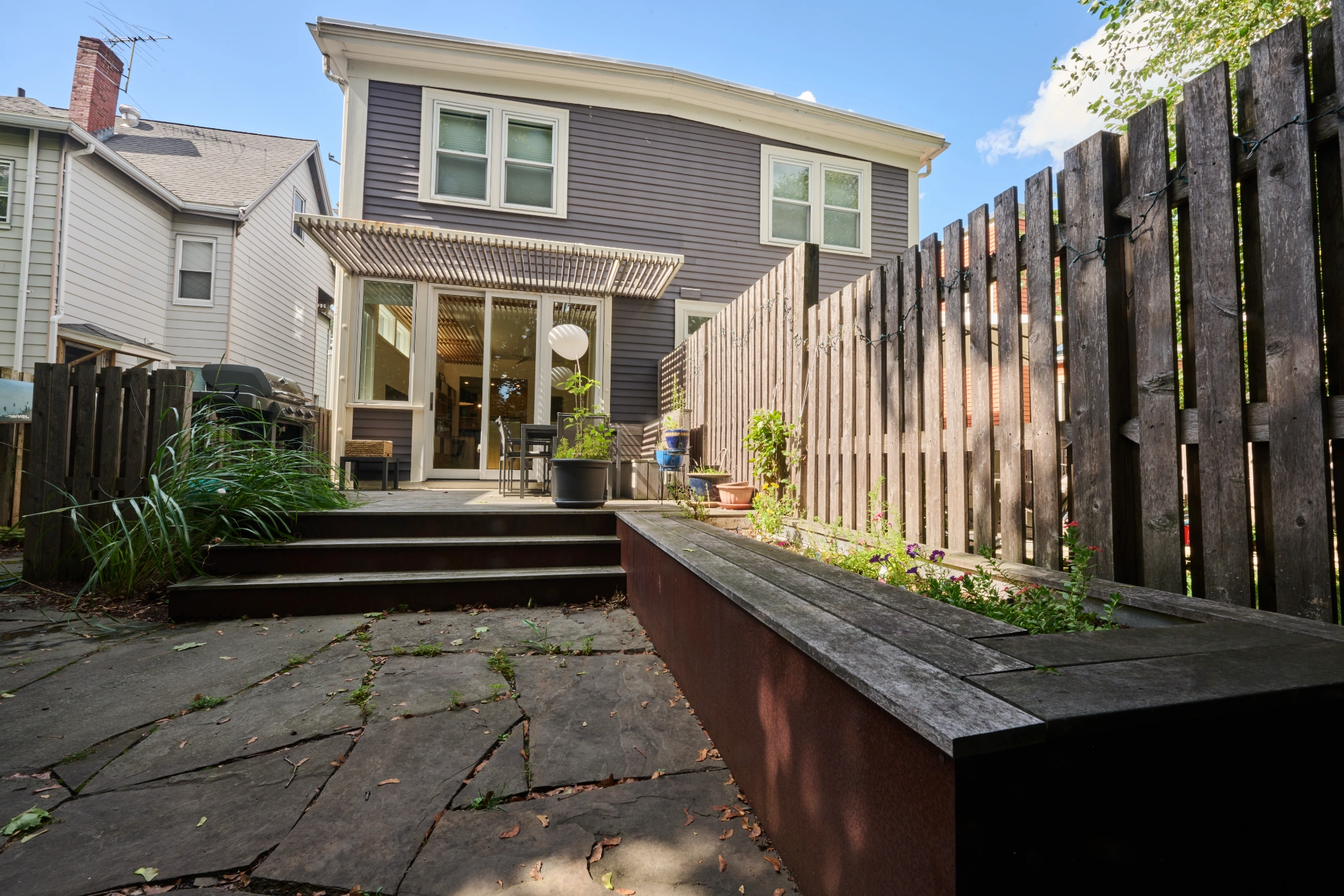 The back of the house from the garden: stone paving, a timber fence and planter, and glass doors under a pergola.