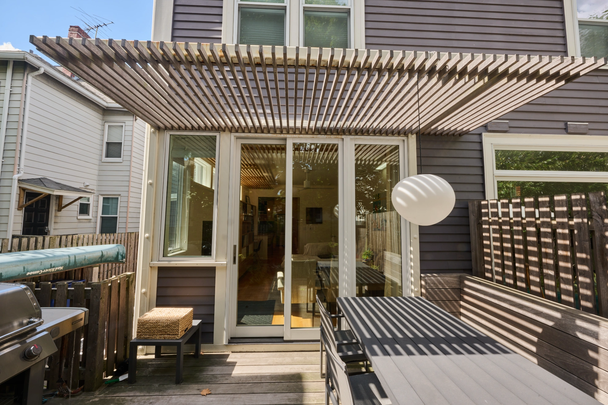 A slatted timber pergola over the deck and the glass doors, with an outdoor table and a paper lantern.