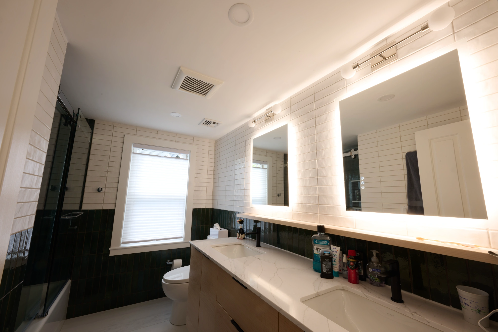 A double vanity under two backlit mirrors, with white tile above a dark green band, and a glass shower beside it.