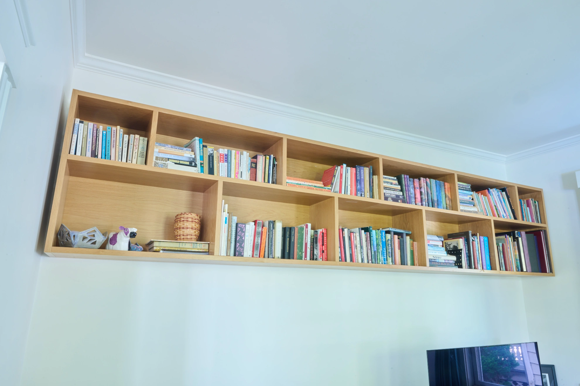 A long maple bookshelf hung high on a white wall, full of books.