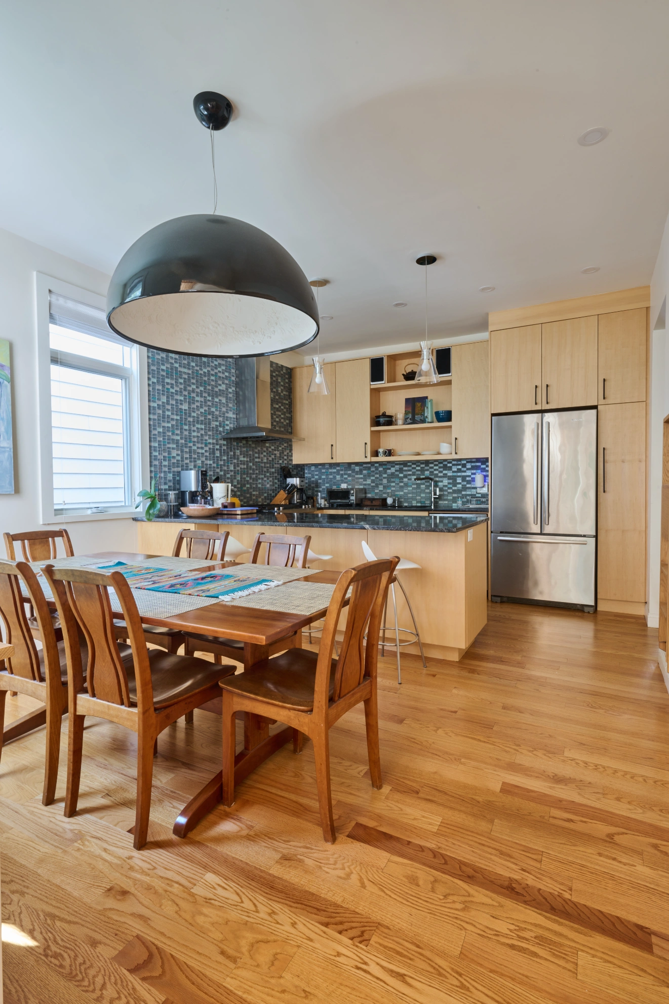 A dining table under a large black dome pendant, with the maple kitchen and a steel refrigerator beyond.