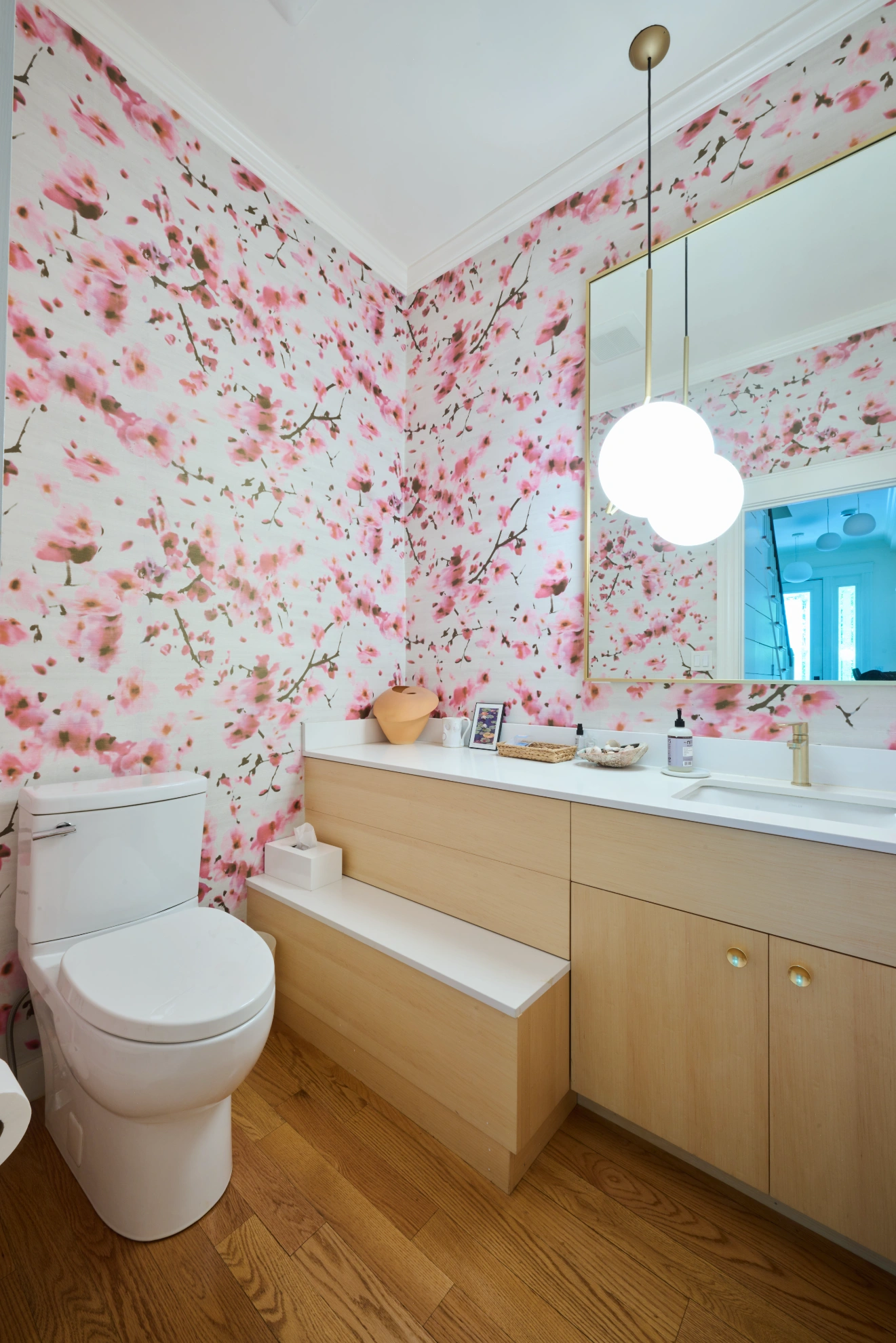 A powder room papered in pink cherry blossom, with a maple vanity and a globe pendant.