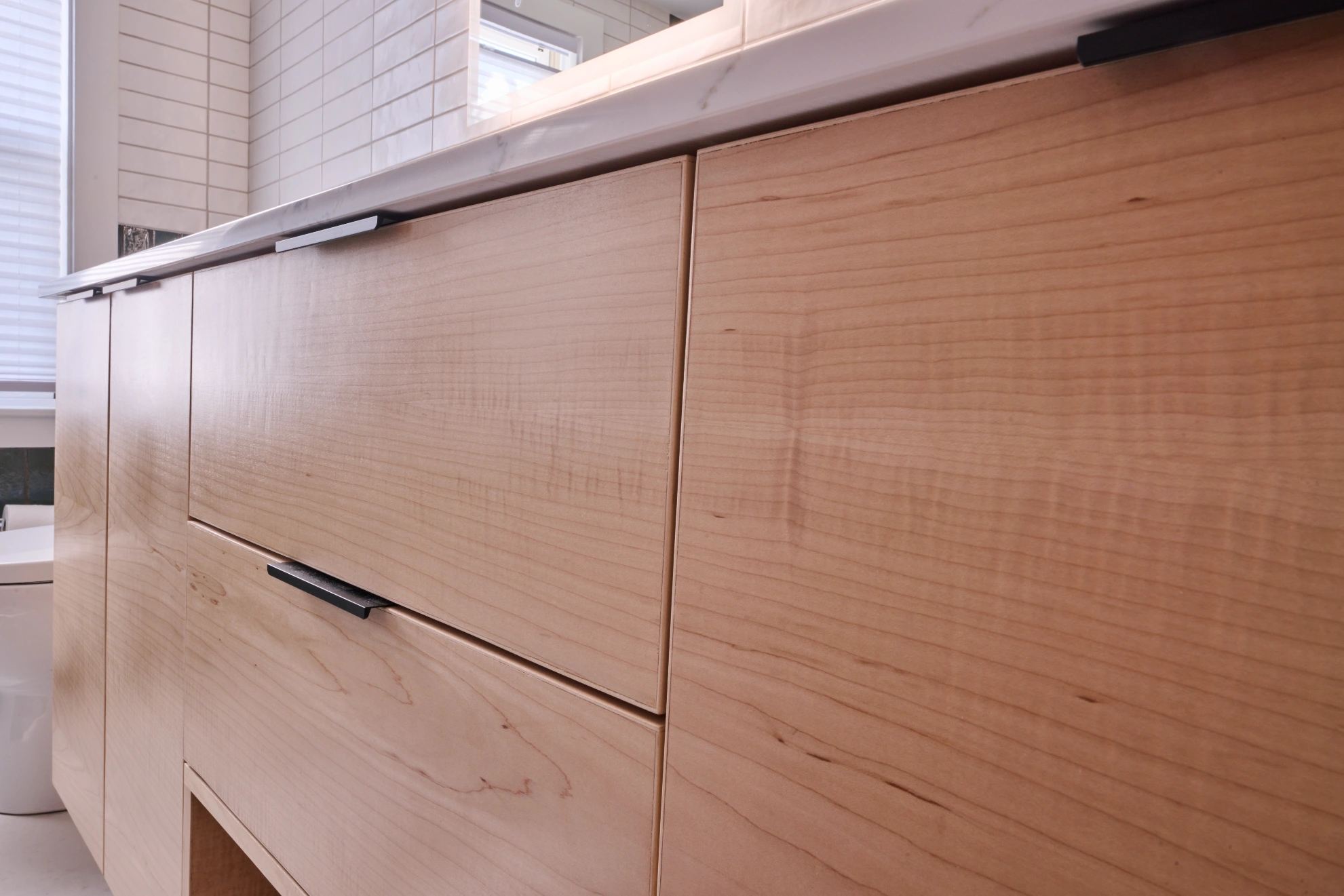 Maple vanity drawers under a white counter, with thin black pulls.