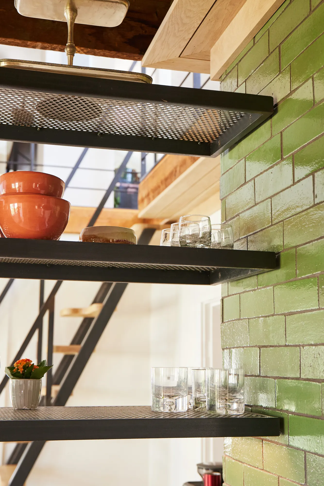 Steel shelves of bowls and glasses against green tile, with the stair behind.