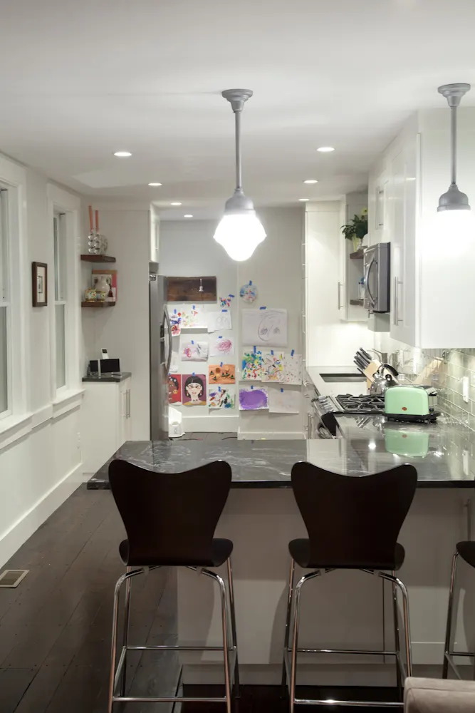 Two black stools at a dark-topped island under pendant lights, the refrigerator beyond covered in children's drawings.