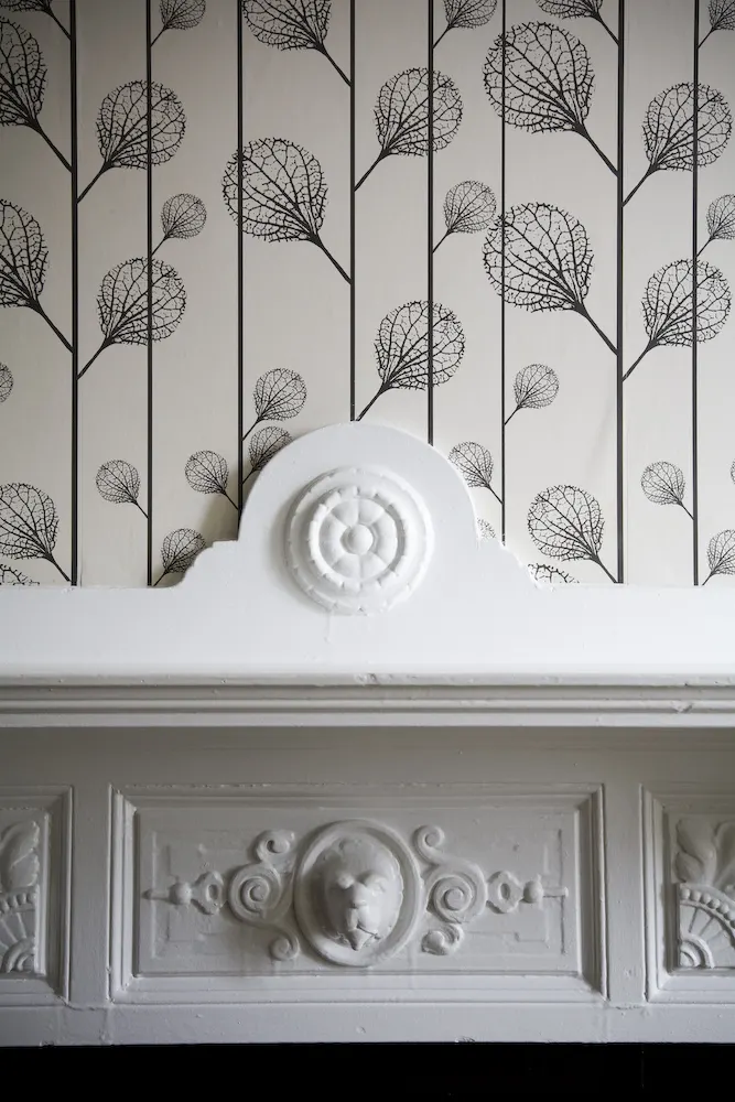 A carved white mantel against black-and-white leaf-pattern wallpaper.