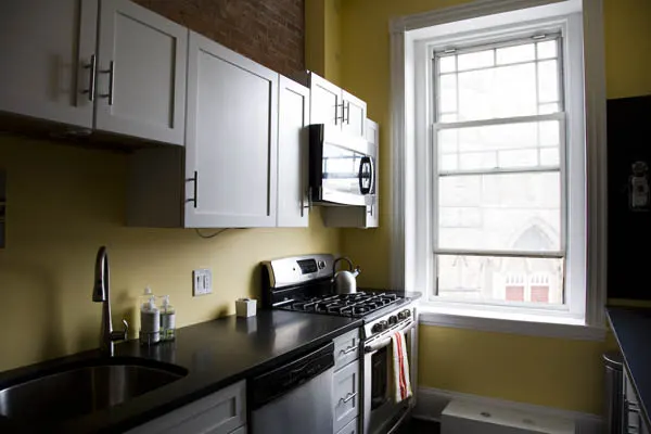 White upper cabinets, dark counters and a steel range by a window, against yellow walls.